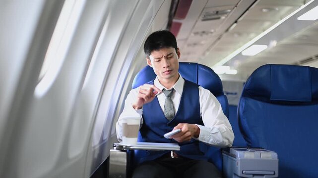 A young Asian executive experiences stress and a headache after checking his phone in business class. Corporate travel fatigue, burnout, and bad news on a flight.