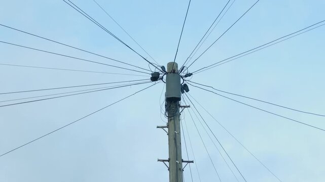 Wooden utility pole with overhead cables against blue sky. Telecommunications junction box and tangled wiring mounted near top of pole. Multiple suspended wires connected to roadside cable
