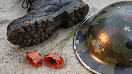 Naklejka premium Displaying dented camouflage helmet on right, muddy combat boot and poppies on burlap, copy space
