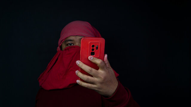 An Asian male wearing a deep maroon long-sleeved shirt has his head and lower face concealed by a vibrant red cloth, leaving only the eyes exposed. The person holds a matching red