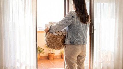 Fototapeta premium Woman carrying laundry basket full of fresh clean laundry, standing on home balcony with warm natural light