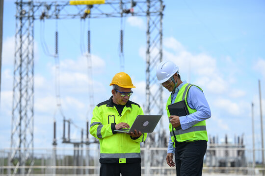 Electrical engineer and technician inspecting substation equipment with laptop and tablet under blue sky, expressing focused teamwork and safety awareness in industrial setting