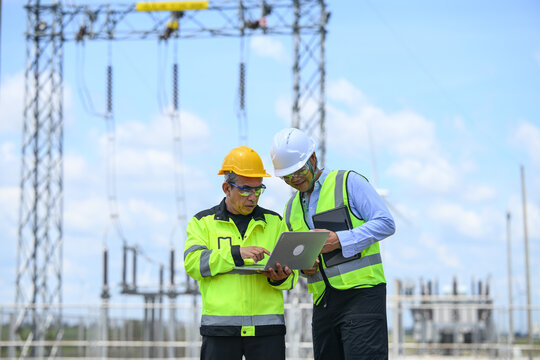 Electrical engineer and technician reviewing laptop data at outdoor power substation during safety inspection, with transmission equipment and blue sky in background
