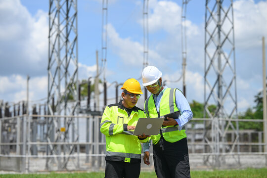 Electrical engineer and technician reviewing laptop data at outdoor power substation, showing focused teamwork during utility infrastructure inspection beneath cloudy blue sky