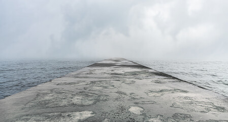 Naklejka premium A concrete pier extends into calm waters under a foggy and overcast sky.