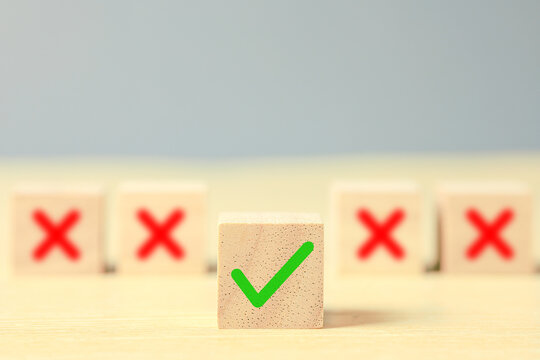 A wooden cube with a green check mark stands in focus between blurred red cross symbols on a bright neutral background, illustrating correct choices, approval, quality, and decisions.