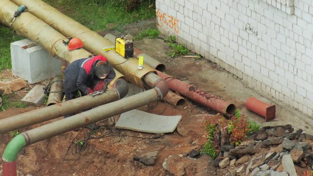 Worker performs arc welding operation on large diameter metal heating pipe, generating bright sparks and smoke during industrial repair project, essential infrastructure maintenance