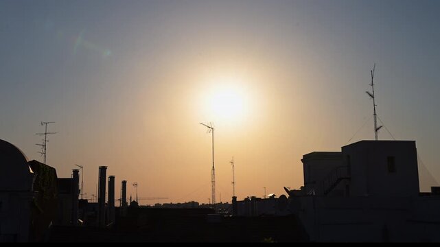 Rooftop perspective captures the bright setting sun over Madrid building silhouettes and communication antennas during the early onset of the historic August 12, 2026 total solar eclipse event, Spain.