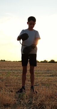 Young boy puts on boxing gloves in open field at sunset, preparing for sparring. Warm golden light highlights concentration, readiness and anticipation before playful combat practice outdoors
