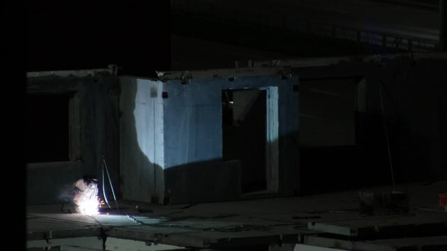Worker performs welding operation at night, generating bright sparks and light during construction of bridge structure or overpass