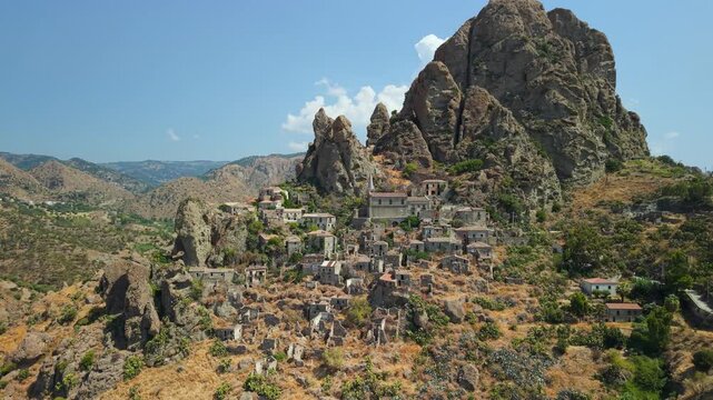 Pentedattilo ghost town in Calabria Italy. Drone side pass reveals the deserted stone village beneath the five-fingered cliffs of Mount Calvario, surrounded by dry fields and rolling hills.