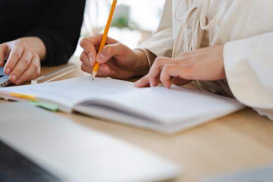 Two colleagues collaborate at a wooden desk during a work meeting. One person holds a yellow pencil, carefully writing notes in an open notebook next to a laptop computer.
