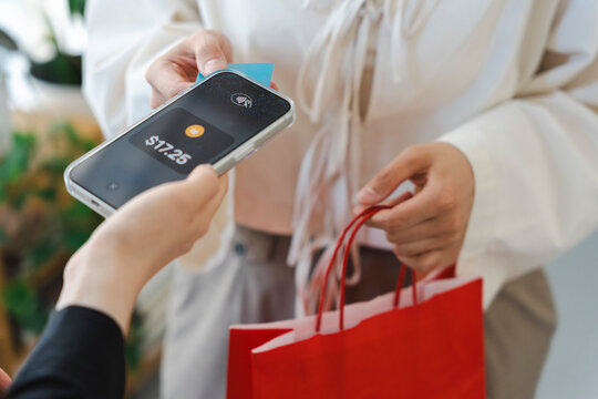 A customer makes a contactless payment using a credit card on a smartphone held by a store employee. The buyer is holding a red shopping bag after a successful purchase.