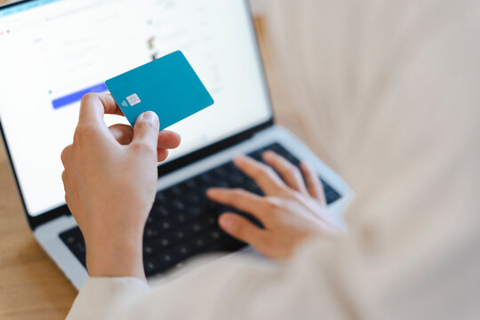 A person sitting at a desk holds a blue payment card in one hand while using a laptop computer to enter credit card details for a secure online shopping transaction.