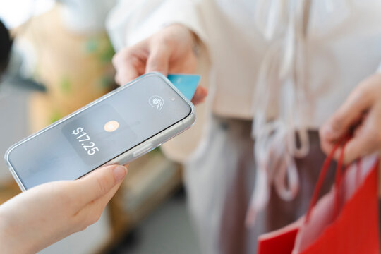A female customer holding shopping bags makes a contactless payment with her credit card by tapping it against a smartphone POS terminal held by a store employee.