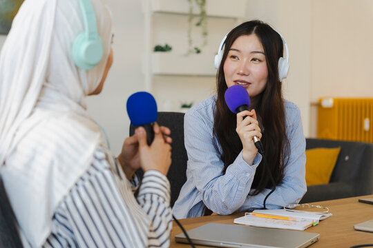 Two diverse women wearing headphones sit together at a wooden desk in a bright office, recording an audio podcast while holding microphones and talking.
