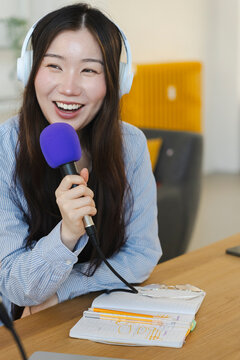 An Asian female content creator wearing white headphones and smiling happily while holding a purple microphone to record a podcast episode at a wooden desk.