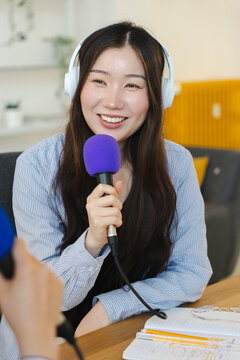 A young Asian woman wearing headphones smiles warmly while holding a purple microphone, actively recording a podcast episode at a wooden table in a bright modern room.