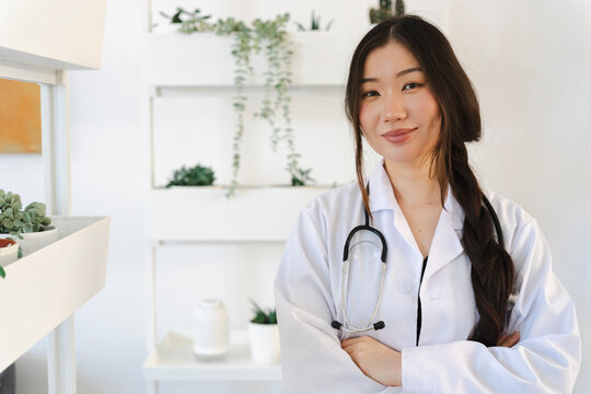 A young female doctor stands in a bright medical office with arms crossed. She wears a white coat and a stethoscope around her neck, smiling gently at the camera.