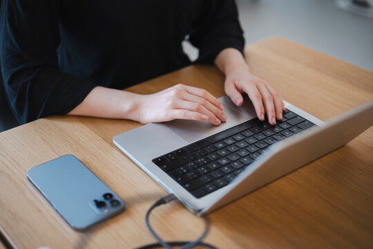 A woman wearing a black shirt sits at a wooden desk and types on a modern laptop computer. A smartphone rests on the table next to the device while she works remotely.