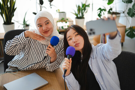 Two young women holding blue microphones pose together for a smartphone selfie while recording a podcast in a bright studio filled with green indoor plants.