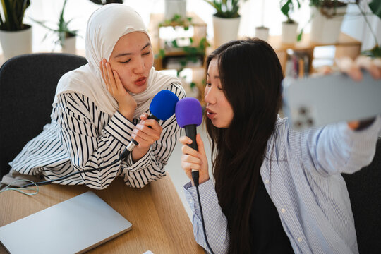 Two young women holding microphones while taking a selfie video with a smartphone. They are recording a podcast or singing together in a modern bright office setting.
