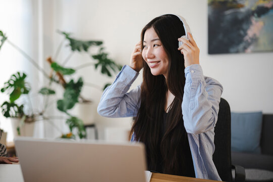 A young Asian woman wearing a blue striped shirt sits at her office desk with a laptop while putting on white headphones and smiling during her workday at home.