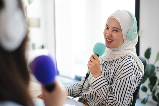 A smiling young woman wearing a white hijab and blue headphones speaks into a microphone during a professional podcast recording session inside a modern broadcasting studio.