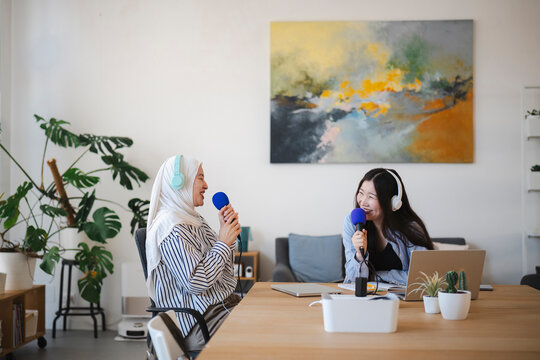 Two smiling young women wearing headphones and holding microphones sit together at a wooden table in a bright room, recording a podcast or interview for their online channel.