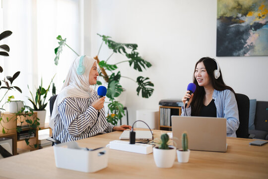 Two young women are recording a podcast in a bright modern studio. They wear headphones and hold microphones while talking to each other across a wooden table with laptops.
