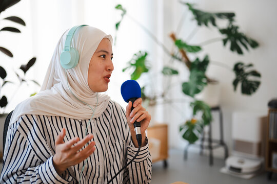 A young Muslim woman wearing a white hijab and teal headphones speaks passionately into a blue microphone while recording a podcast inside a bright room with indoor plants.