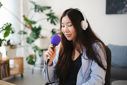 A young Asian woman with long dark hair wears headphones and speaks into a microphone. She records a podcast or audio broadcast in a bright room with indoor plants.