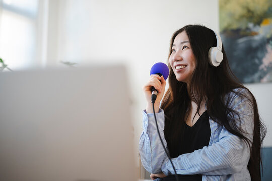 A young Asian woman wearing white headphones smiles while holding a blue microphone in her hand, recording audio for a podcast or singing at home during the day.