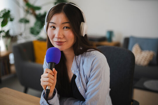 An Asian woman wearing headphones and holding a purple microphone while recording an audio broadcast at a desk. She looks forward with a gentle and friendly smile.