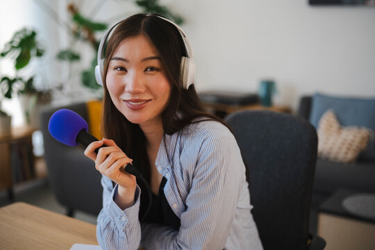 A young Asian woman wearing headphones smiles while holding a microphone in her home office. She is recording an audio podcast or speaking during a live online broadcast.
