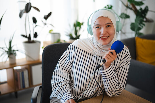 A young Muslim woman wearing a hijab and headphones holds a microphone while recording a podcast in her cozy home studio. She smiles warmly at the camera.