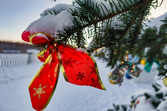 A decorated tree with red ribbons and ornaments sits in the snow. The setting shows a winter landscape with a fence in the background and lights