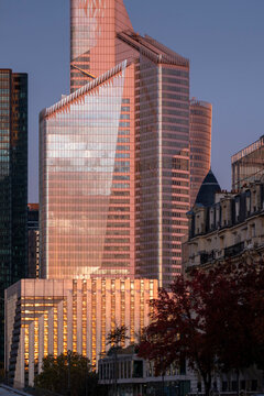 Warm sunset light gives a glass skyscraper facade a copper glow above the city in La Defense near Paris France with surrounding buildings visible in shadow