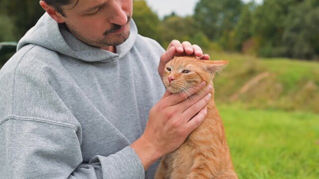 A close-up shot of a man affectionately petting a ginger cat on the lawn. The owner gently kisses his pet while sitting outdoors during the day.
