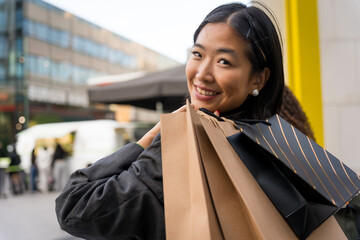 close up portrait of young smiling asian woman in her 30s with shopping bags watch happy and...