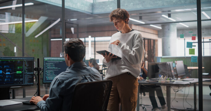 Caucasian Female Project Manager With Tablet Talking to South Asian Male Software Engineer Working Using a Computer With Two Monitors Showing Program Code During a Meeting in a Modern Startup Office