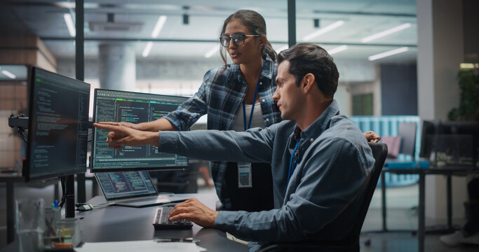 Caucasian Male Backend Developer Pointing at Monitor With Software Code During Meeting With South Asian Female Data Engineer in Modern Tech Office, Talking and Working Using Computer