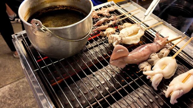 Close up shot of street food vendor hands using metal tongs to flip and arrange fresh squid skewers on a hot grill rack next to a sauce pot at a market.
