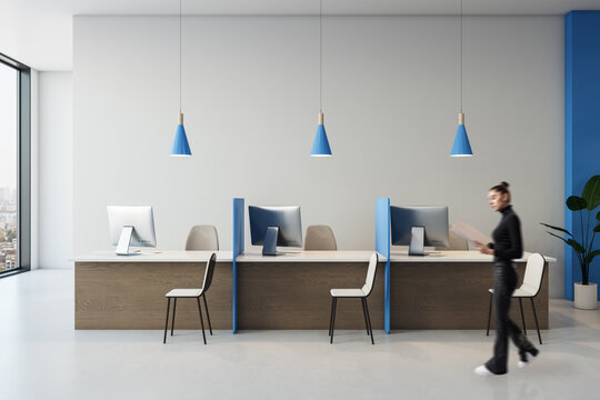 Modern bank reception interior with businesswoman walking past service desks, computers, pendant lights, and blue partitions in bright corporate office.