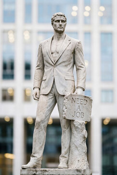 A statue of David dressed in a business suit, holding a sign that reads "Need a job," set against the backdrop of a modern city with skyscrapers