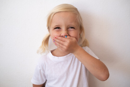 Cute Little Blonde Girl Covering Mouth with Hand and Smiling on White Background