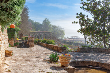 Naklejka premium A sunny stone terrace surrounded by green trees, blooming flowers, and historic architecture overlooking a scenic view.