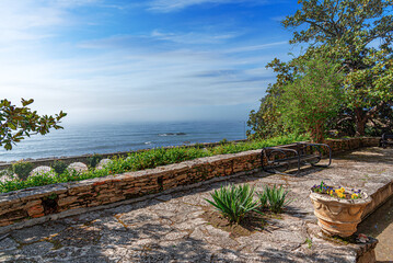 Naklejka premium A sunny stone terrace surrounded by lush greenery and flowers overlooks a calm sea under a clear blue sky.