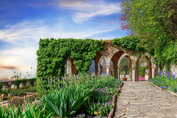 Naklejka premium A beautiful stone pathway leads through a sunny garden with lush green arches, colorful flowers, and a clear blue sky.