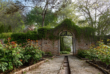 Naklejka premium An arched stone wall covered in green ivy leads into a peaceful garden surrounded by blooming roses and tall trees.